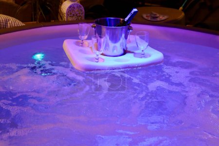 Téléchargez les photos : Bouteille de champagne dans un seau à glace et 4 verres sur un flotteur dans un bain jacuzzi bouillonnant avec des lumières violettes sous l'eau - en image libre de droit