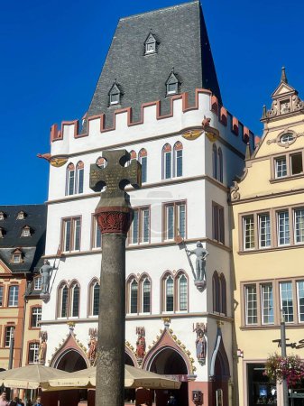 Photo for Oldest Market cross in Germany, with Latin dedication from year 958. Gothic Steipe house with figures of saints and knights behind it. Trier, Rhineland-Palatinate, Germany - September 14, 2023 - Royalty Free Image