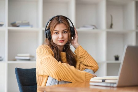 Asian woman in headphones listening to music happily on her laptop enjoying listening to music during work during a relaxing break from studying online