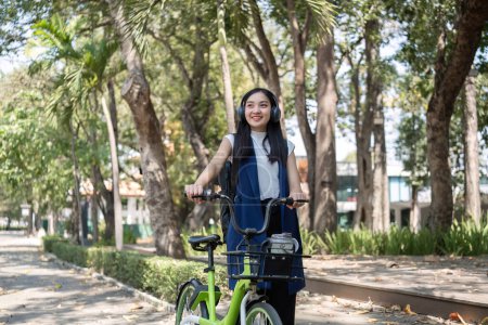 Foto de Una joven asiática viaja al trabajo en bicicleta por una ciudad verde, llevando una mochila y usando una taza de bebida reutilizable para evitar dañar el medio ambiente.. - Imagen libre de derechos