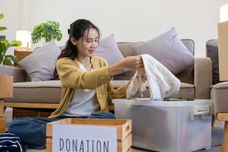 Cute Asian woman sitting next to sofa in living room sorting unwanted clothes for donation..