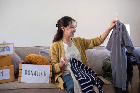 Cute Asian woman sits on a sofa sorting unwanted clothes to donate..