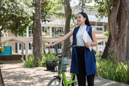 Foto de Una joven asiática viaja al trabajo en bicicleta por una ciudad verde, llevando una mochila y usando una taza de bebida reutilizable para evitar dañar el medio ambiente.. - Imagen libre de derechos