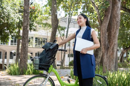 Eine junge Asiatin pendelt mit dem Fahrrad in einer grünen Stadt zur Arbeit, trägt einen Rucksack und verwendet einen wiederverwendbaren Trinkbecher, um der Umwelt nicht zu schaden..