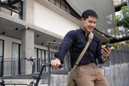 Ein junger asiatischer Geschäftsmann fährt mit dem Fahrrad zur Arbeit. Draußen vor dem Büro stehen, telefonieren und über Geschäfte reden. Konzept zur Verringerung von Energie und Luftverschmutzung..