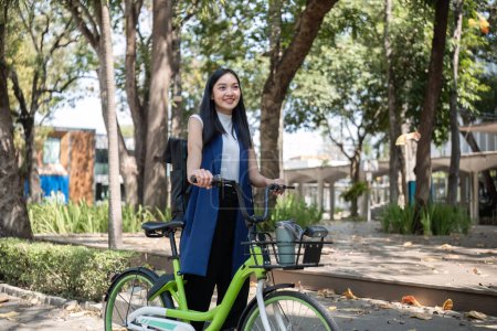 Eine junge Asiatin pendelt mit dem Fahrrad in einer grünen Stadt zur Arbeit, trägt einen Rucksack und verwendet einen wiederverwendbaren Trinkbecher, um der Umwelt nicht zu schaden..