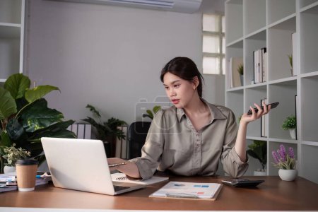 Junge asiatische Geschäftsfrau sitzt am Telefon in einem Online-Geschäftstreffen mit einem Laptop in einem modernen Homeoffice, das mit schattigen grünen Pflanzen dekoriert ist..