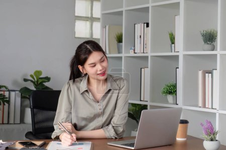 Asian businesswoman or accountant happily sitting and working with laptop on finance and business administration. On the desk in the office decorated with green plants..