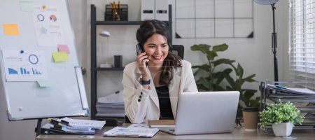 Junge asiatische Geschäftsfrau sitzt am Telefon in einem Online-Geschäftstreffen mit einem Laptop in einem modernen Homeoffice, das mit schattigen grünen Pflanzen dekoriert ist..