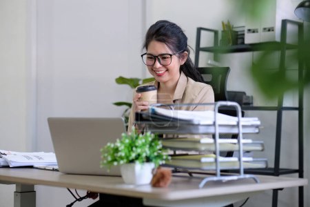 Asiatische Geschäftsfrau oder Buchhalterin, die fröhlich sitzt und mit Laptop an Finanzen und Betriebswirtschaft arbeitet. Auf dem Schreibtisch im Büro mit grünen Pflanzen dekoriert..