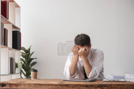 Joven hombre de negocios sentado en un escritorio con la cabeza en las manos, mostrando decepción y frustración en el trabajo. Ambiente de oficina con decoración moderna.