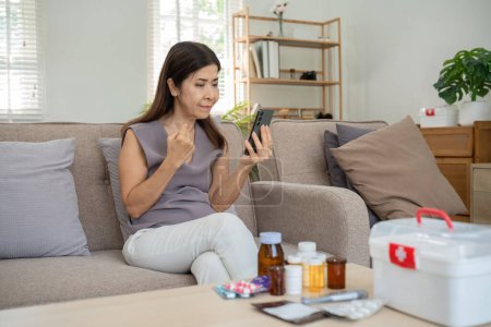 Ältere Frau, die Medikamente in der Hand hält, während sie auf einem Smartphone berät, sitzt in einem modernen Wohnzimmer mit Medikamenten auf dem Tisch.