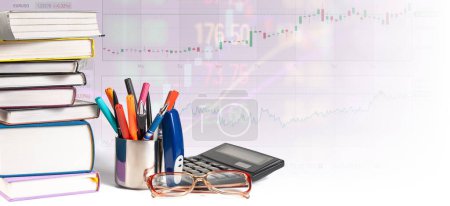 A collection of books is piled next to office supplies including pens, calculator, and glasses, all set against the backdrop of an illuminated stock market graph.