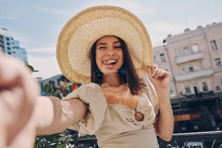 Téléchargez les photos : Jeune femme ludique en chapeau élégant faisant selfie tout en se relaxant sur le balcon - en image libre de droit