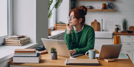 Téléchargez les photos : Jeune rousse femme regardant réfléchi tout en travaillant sur ordinateur portable au comptoir de la cuisine - en image libre de droit