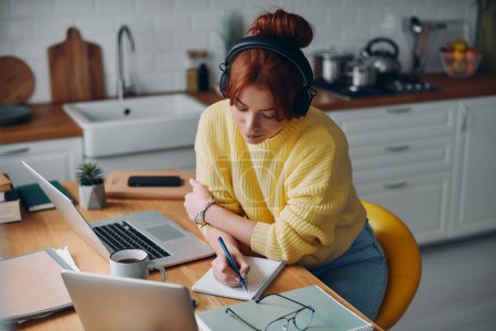 Photo for Concentrated woman in headphones making notes in her pad while sitting at the kitchen counter - Royalty Free Image