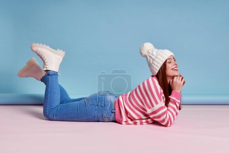 Téléchargez les photos : Femme rêveuse en vêtements d'hiver gardant les yeux fermés et souriant tout en étant couché sur fond bleu - en image libre de droit