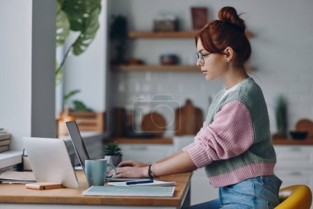 Téléchargez les photos : Jeune rousse femme travaillant sur ordinateur portable tout en étant assis au comptoir de la cuisine à la maison - en image libre de droit