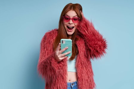 Überraschte junge Frau im flauschigen Mantel blickt auf ihr Smartphone vor blauem Hintergrund