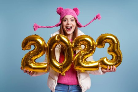 Photo for Excited woman in winter coat holding gold colored numbers and smiling against blue background - Royalty Free Image