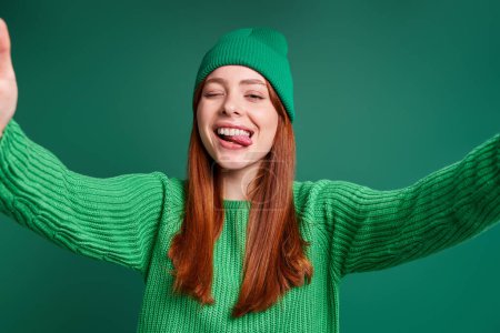 Téléchargez les photos : Belle jeune femme faisant selfie et grimacant tout en se tenant debout sur fond vert - en image libre de droit