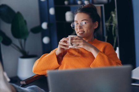 Téléchargez les photos : Jeune femme détendue dégustant un café tout en étant assise à son lieu de travail au bureau - en image libre de droit