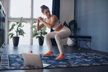 Foto de Mujer joven y segura en ropa deportiva de pie en posición de okupa mientras hace ejercicio en casa - Imagen libre de derechos