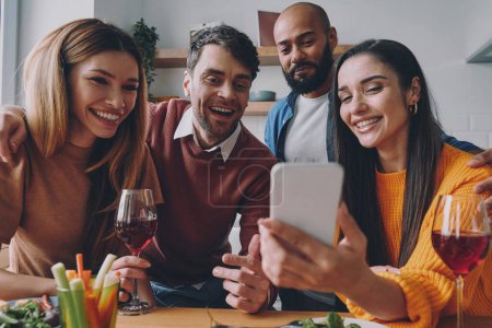 Foto de Grupo de jóvenes mirando el teléfono inteligente y sonriendo mientras disfrutan del tiempo juntos - Imagen libre de derechos