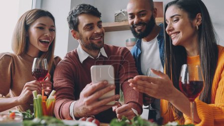 Téléchargez les photos : Groupe de jeunes gens heureux discutant de quelque chose tout en regardant le téléphone intelligent ensemble - en image libre de droit