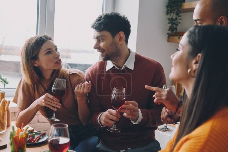 Foto de Jóvenes felices bebiendo vino y hablando mientras disfrutan de la cena en casa juntos - Imagen libre de derechos