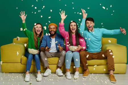 Foto de Grupo de jóvenes felices lanzando palomitas de maíz mientras están sentados en el sofá contra el fondo rosa - Imagen libre de derechos