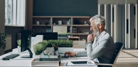 Téléchargez les photos : Homme d'affaires mature concentré travaillant sur ordinateur portable tout en étant assis à son lieu de travail dans le bureau - en image libre de droit