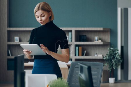 Téléchargez les photos : Jeune femme d'affaires utilisant une tablette numérique tout en se tenant près de son lieu de travail au bureau - en image libre de droit