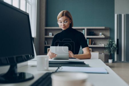 Téléchargez les photos : Confiant jeune femme d'affaires en utilisant une tablette numérique tout en travaillant au bureau - en image libre de droit