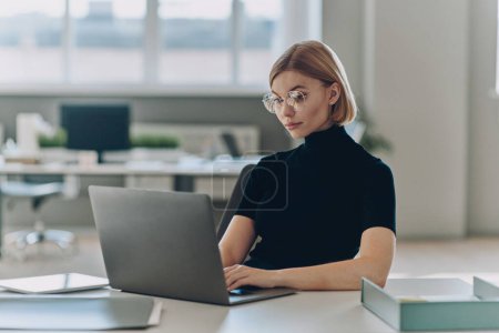 Téléchargez les photos : Confiant jeune femme d'affaires utilisant un ordinateur portable tout en étant assis à son lieu de travail dans le bureau - en image libre de droit