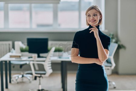 Foto de Mujer de negocios confiada mirando hacia otro lado y sonriendo mientras está de pie en la oficina - Imagen libre de derechos