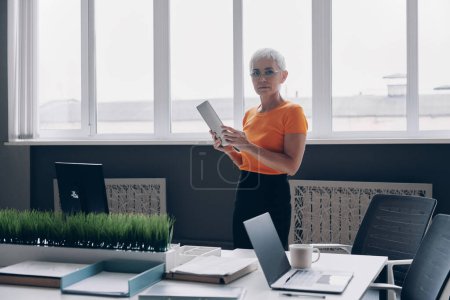 Téléchargez les photos : Femme d'affaires senior confiante tenant une tablette numérique tout en travaillant dans le bureau - en image libre de droit