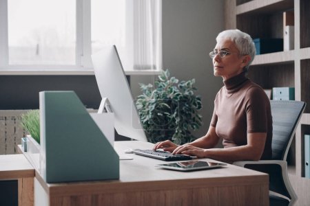 Foto de Empresa senior de confianza que usa la computadora mientras está sentada en su lugar de trabajo en la oficina - Imagen libre de derechos