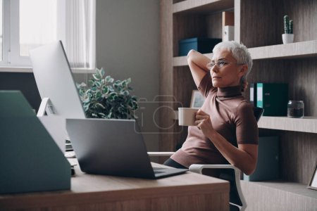 Foto de Relajada mujer de negocios senior disfrutando de una bebida caliente mientras está sentada en su lugar de trabajo en la oficina - Imagen libre de derechos
