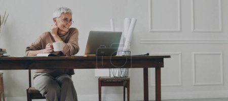 Téléchargez les photos : Heureuse femme d'affaires âgée profitant d'une boisson chaude tout en utilisant un ordinateur portable à son lieu de travail - en image libre de droit