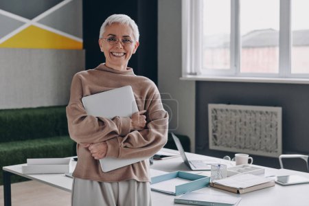 Téléchargez les photos : Femme d'affaires senior tenant un ordinateur portable et souriant tout en se tenant près de son lieu de travail - en image libre de droit