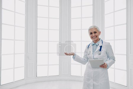 Téléchargez les photos : Heureuse femme médecin tenant tablette numérique et gestuelle tout en se tenant contre une fenêtre - en image libre de droit