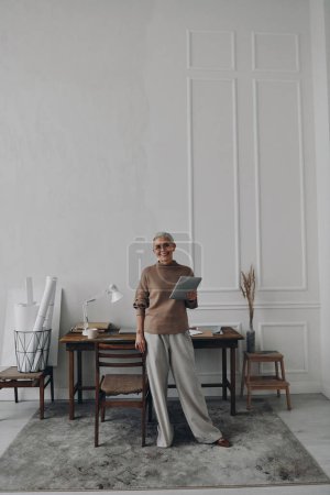 Téléchargez les photos : Heureuse femme âgée tenant tablette numérique tout en se tenant près de son lieu de travail - en image libre de droit