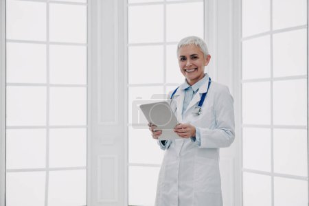 Photo for Confident female doctor holding digital tablet and smiling while standing against a window - Royalty Free Image