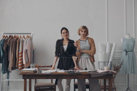 Foto de Dos confiadas diseñadoras de moda mirando a la cámara mientras están de pie en el taller juntas - Imagen libre de derechos