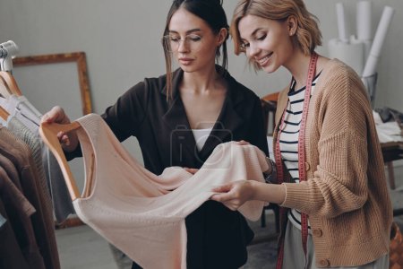 Téléchargez les photos : Deux créatrices de mode confiantes examinant des vêtements tout en travaillant en atelier - en image libre de droit