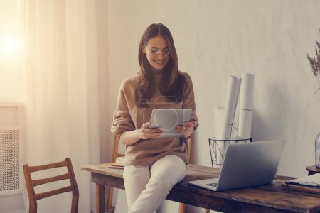 Téléchargez les photos : Joyeux jeune femme travaillant sur tablette numérique tout en étant assis sur le bureau dans le bureau créatif - en image libre de droit