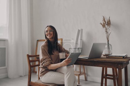 Téléchargez les photos : Joyeux jeune femme tenant tablette numérique tout en étant assis sur le bureau dans le bureau créatif - en image libre de droit