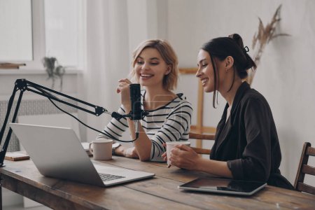 Photo for Two attractive young women talking and smiling while providing live stream in studio together - Royalty Free Image