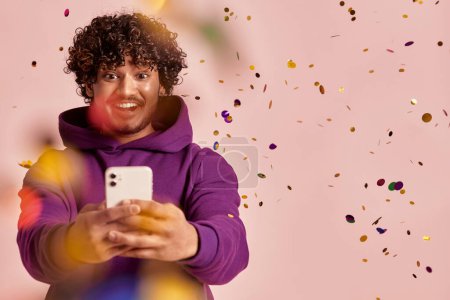 Téléchargez les photos : Homme indien joyeux utilisant un téléphone intelligent tandis que les confettis volent autour sur fond rose - en image libre de droit